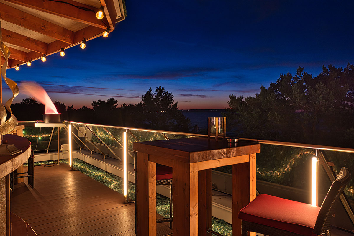 Dachterrasse der Arkona Sky Bar am Abend mit Blick auf die Ostsee, Holztisch, Stuhl und sanfter Sommerbeleuchtung.